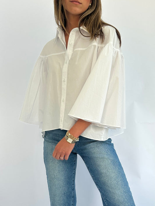 Woman wearing a white button-up shirt with wide, flared sleeves and blue jeans. Her hand rests on her hip, displaying gold bracelets.