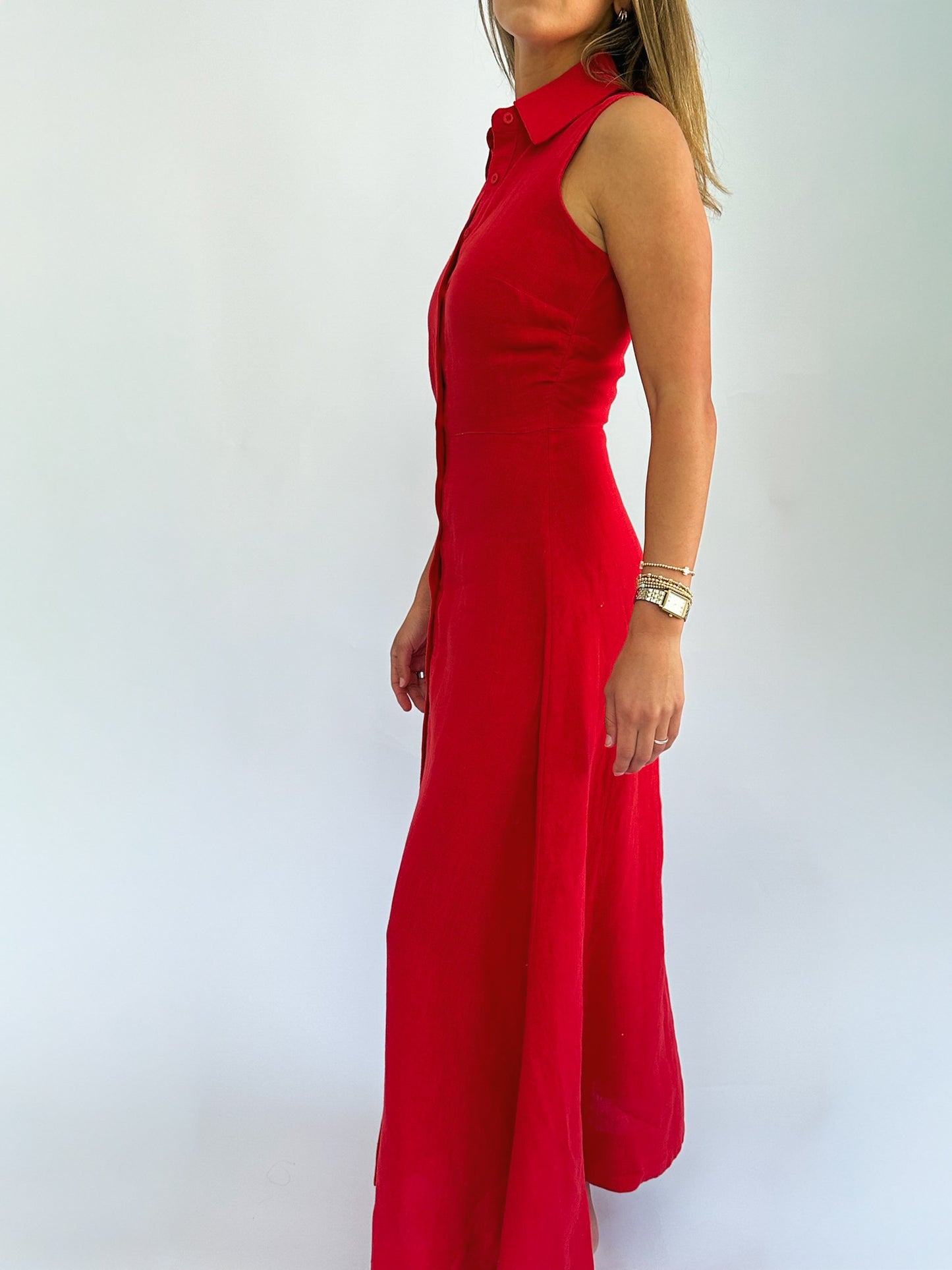 Woman in a sleeveless red dress with a collared top, side view. She wears a gold bracelet, standing against a plain white background.