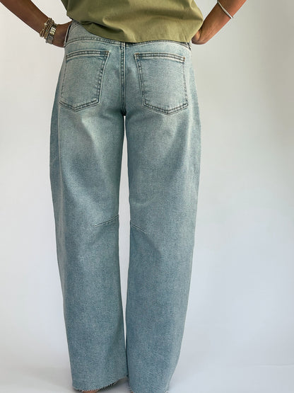 Back view of a person in light blue wide-leg jeans with hands on hips, wearing a green top. The setting is neutral, highlighting casual attire.