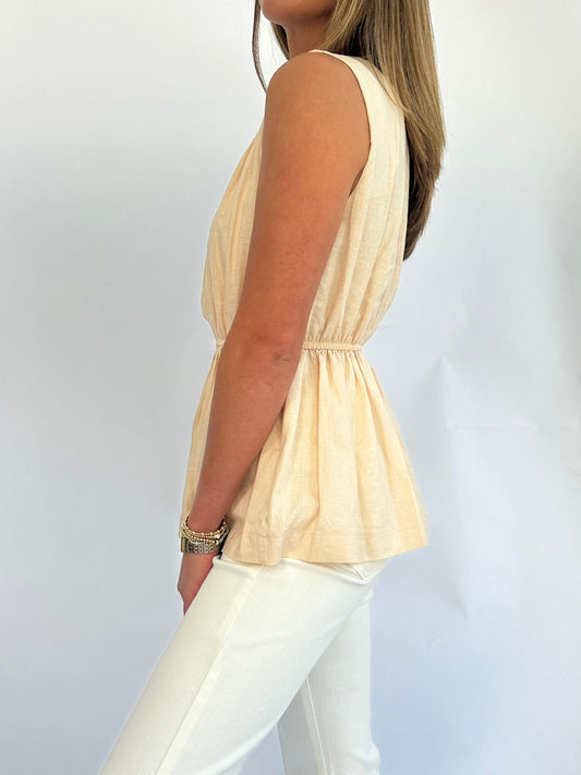 A person wearing a sleeveless, peach-colored, gathered top. The outfit features a cinched waist and is paired with light-colored pants, conveying a casual and elegant style.