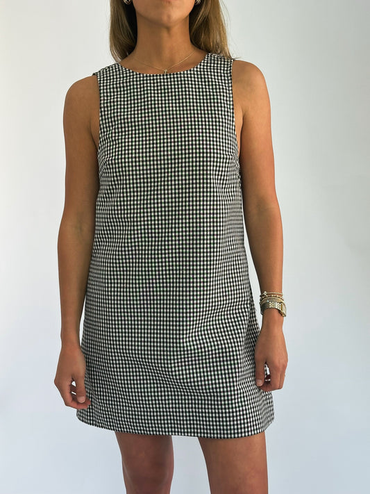 A woman in a sleeveless, black-and-white checkered dress stands confidently. She wears a gold bracelet, conveying a casual yet stylish vibe.