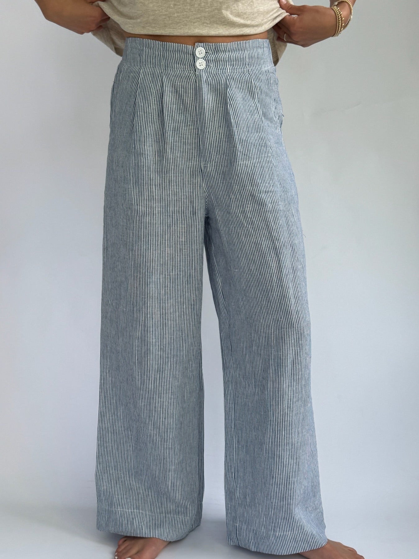 Person wearing high-waisted, blue and white striped, wide-leg linen pants with buttons. Hands on hips; casual, relaxed tone. Barefoot on plain background.