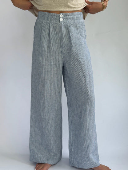 Person wearing high-waisted, blue and white striped, wide-leg linen pants with buttons. Hands on hips; casual, relaxed tone. Barefoot on plain background.