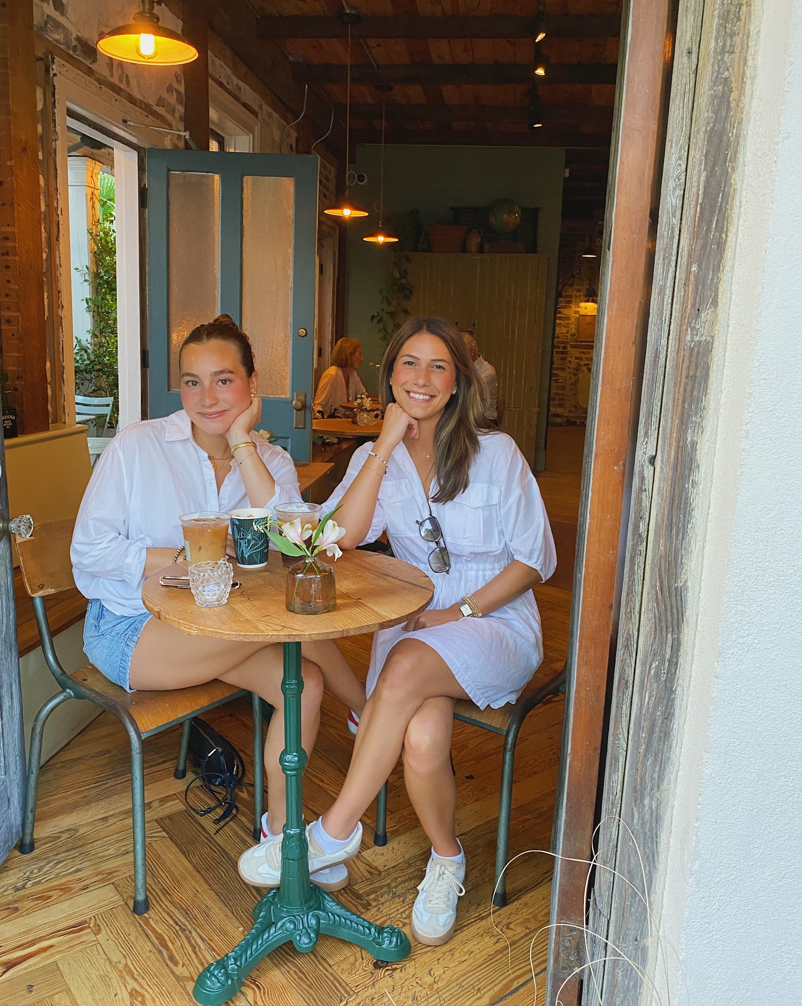 Two women in casual white outfits sit at a café table, smiling. Iced drinks and flowers on the table. Warm, cozy atmosphere.