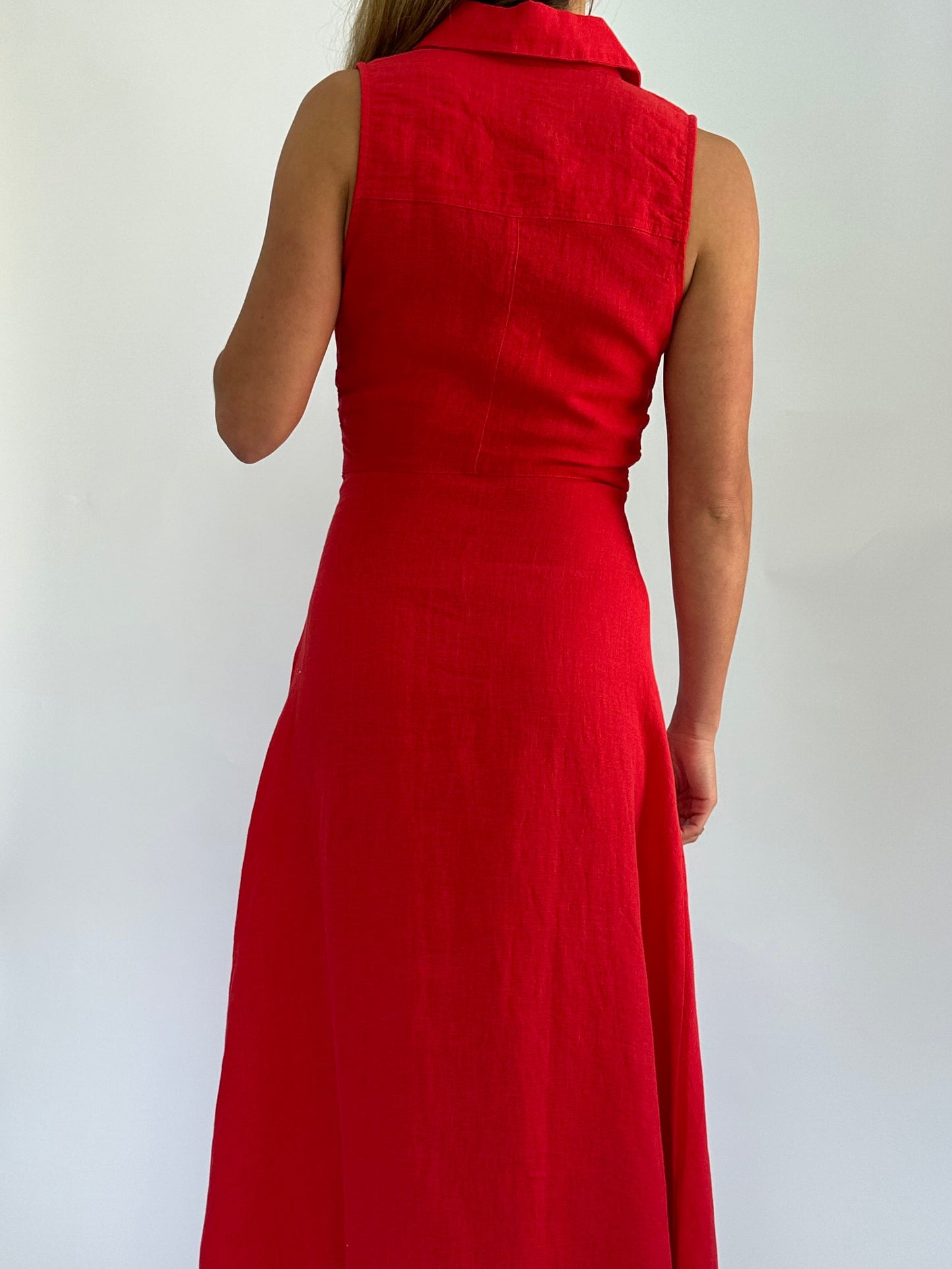 A woman in a sleeveless, knee-length red dress stands with her back to the camera against a plain white background, conveying a simple, elegant style.