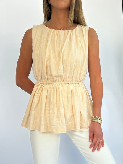 A person wearing a sleeveless, peach-colored, gathered top. The outfit features a cinched waist and is paired with light-colored pants, conveying a casual and elegant style.