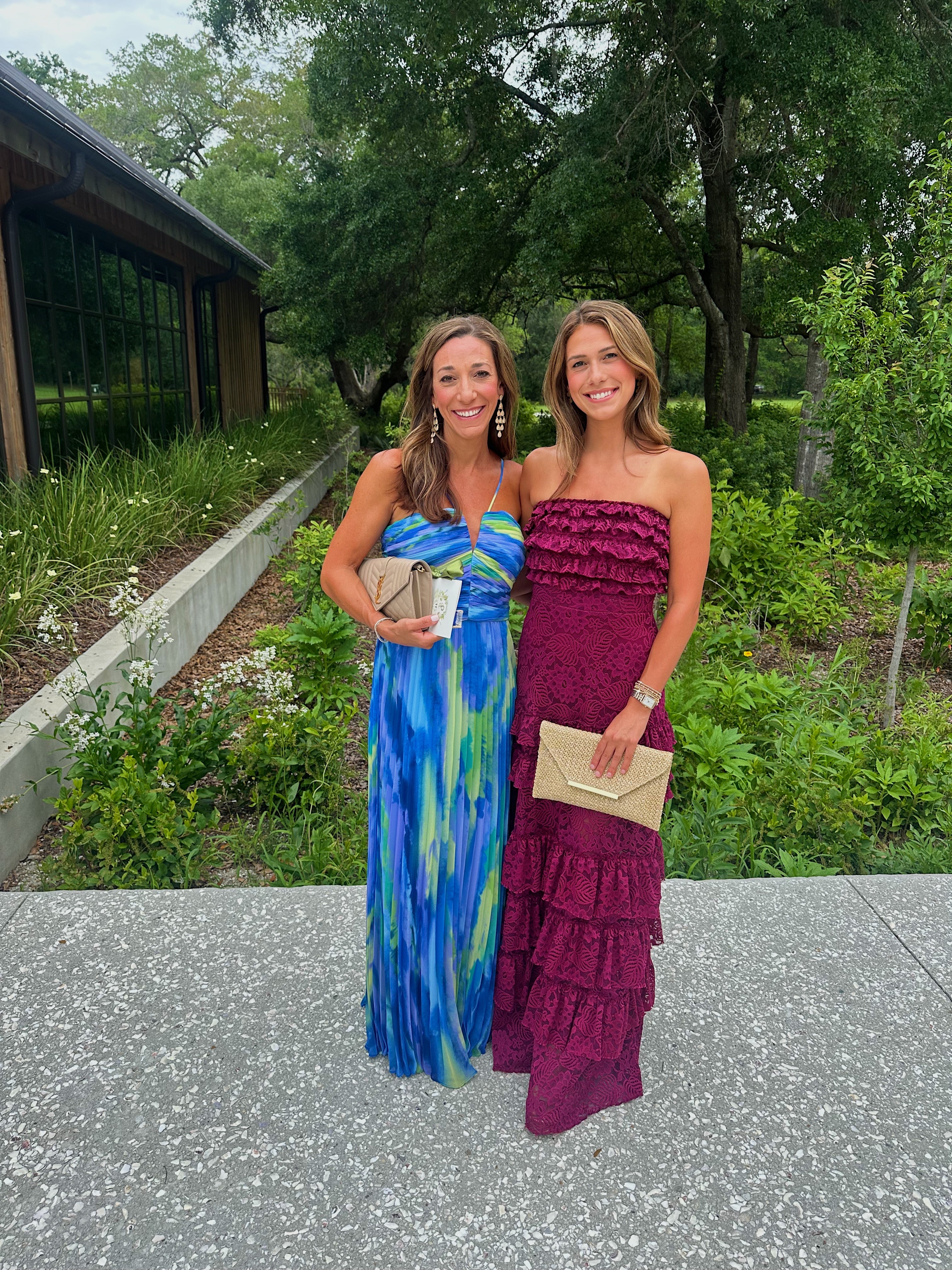 Two women smiling outdoors; one in a blue-green dress, the other in a maroon strapless dress, holding clutches. Lush greenery in the background.
