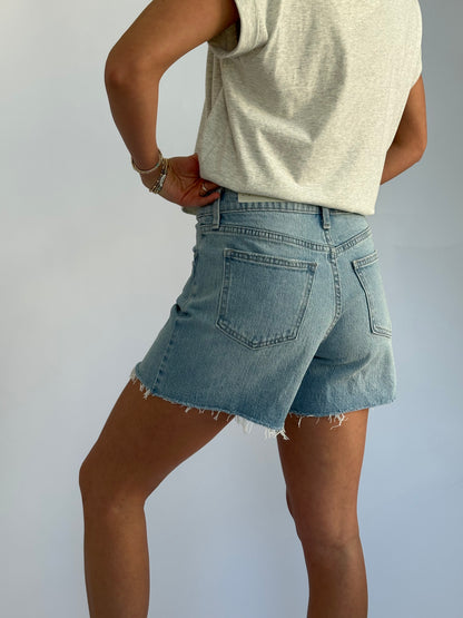 Person wearing a casual gray t-shirt and light blue denim shorts. Hands are on hips, conveying a relaxed and confident vibe against a plain background.