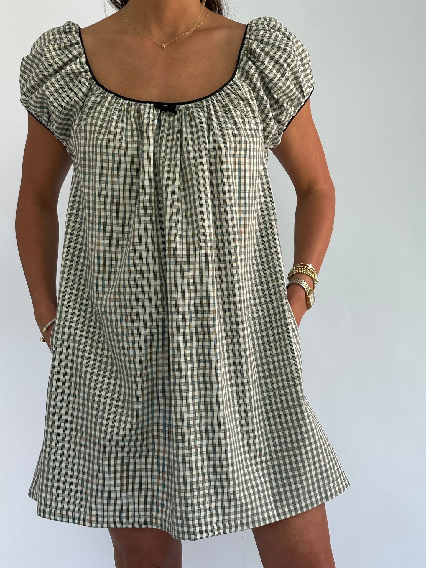 Woman wearing a black-and-white checkered dress with puffed sleeves, hands in pockets. She accessorizes with a gold necklace and bracelets.