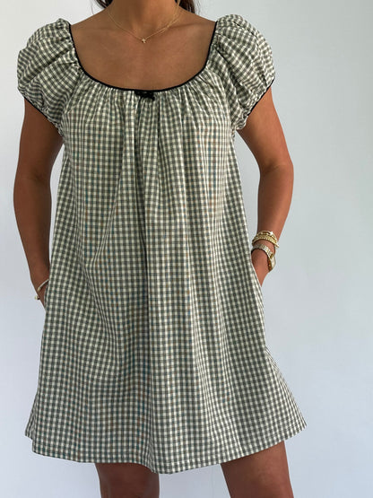 Woman wearing a black-and-white checkered dress with puffed sleeves, hands in pockets. She accessorizes with a gold necklace and bracelets.