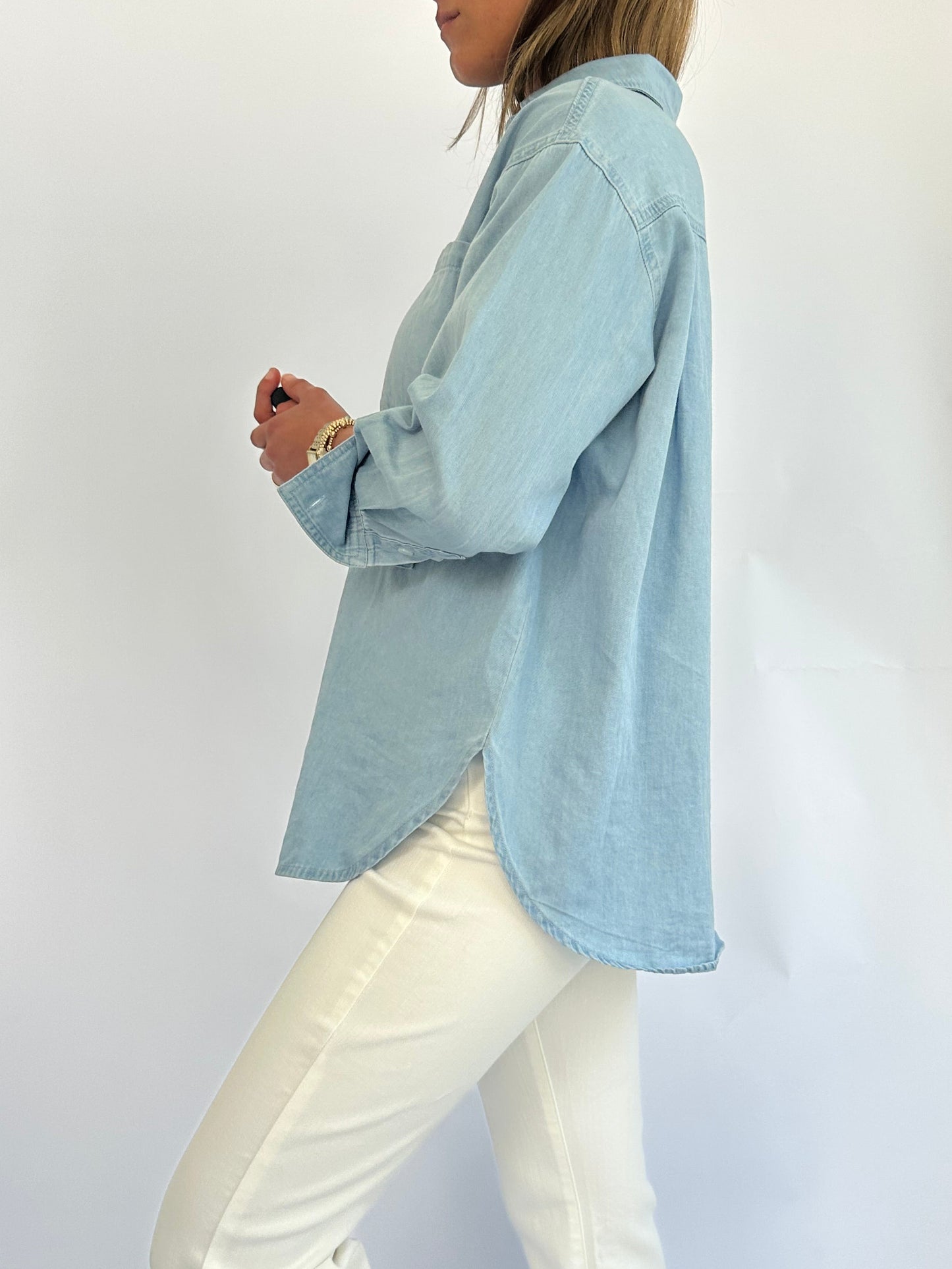 A woman in a light blue denim button-up shirt and white pants is standing sideways against a plain background, exuding a casual, relaxed vibe.