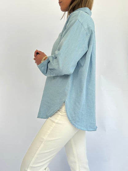 A woman in a light blue denim button-up shirt and white pants is standing sideways against a plain background, exuding a casual, relaxed vibe.