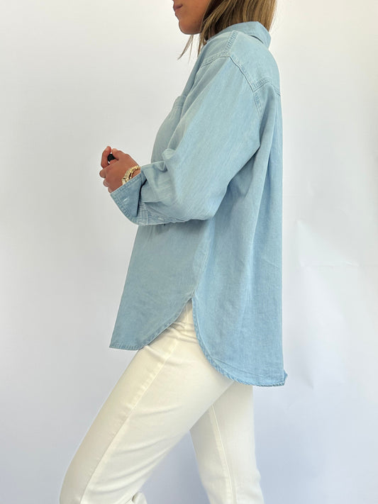 A woman in a light blue denim button-up shirt and white pants is standing sideways against a plain background, exuding a casual, relaxed vibe.