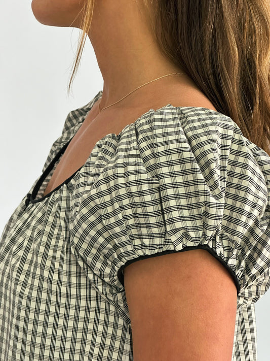 Close-up of a woman in a checkered dress with short puff sleeves, showcasing a delicate gold necklace. The tone is elegant and minimalist.