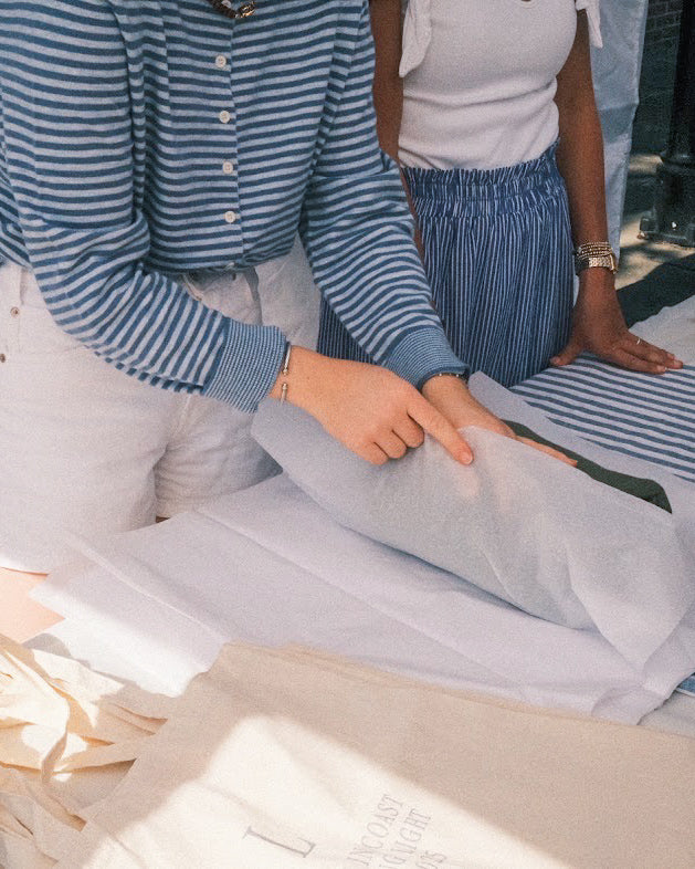 Two people wrapping a shirt on a table outdoors. One wears a blue striped shirt; the other, a white tank with a blue skirt, creating a calm, collaborative scene.