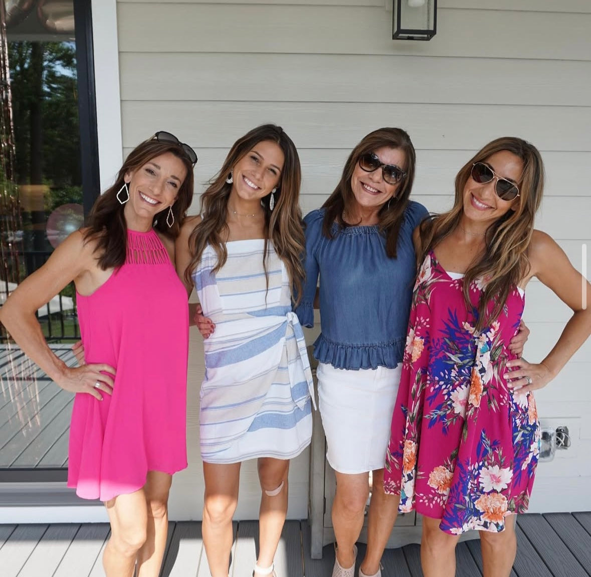 Four women smiling and posing outside. They wear vibrant summer dresses, including pink and floral designs. The mood is joyful and relaxed.