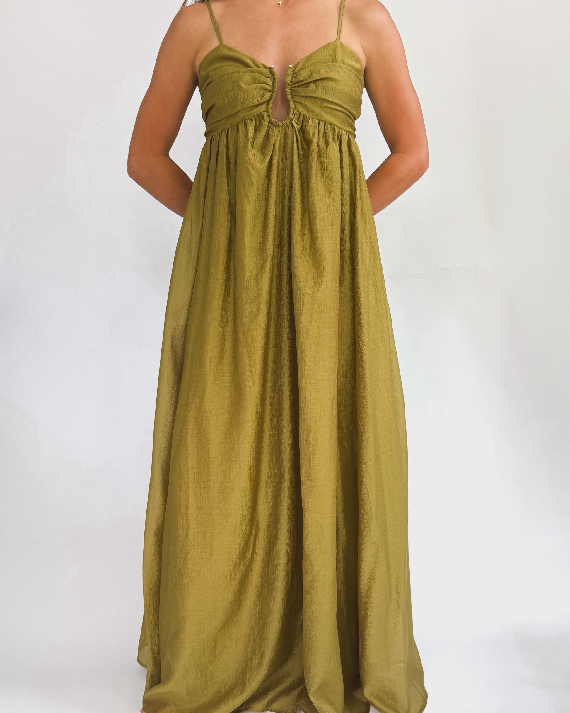 A person wearing a long, olive green dress stands in front of a plain white background. The dress has thin straps and a gathered bust, exuding elegance.
