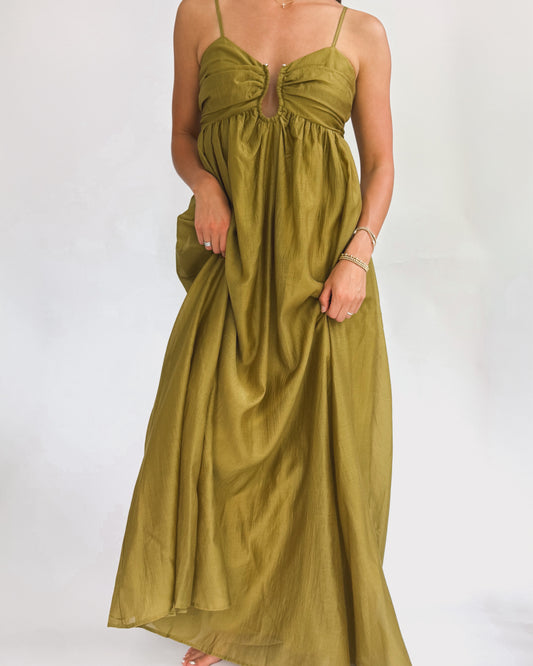 A person wearing a long, olive green dress stands in front of a plain white background. The dress has thin straps and a gathered bust, exuding elegance.