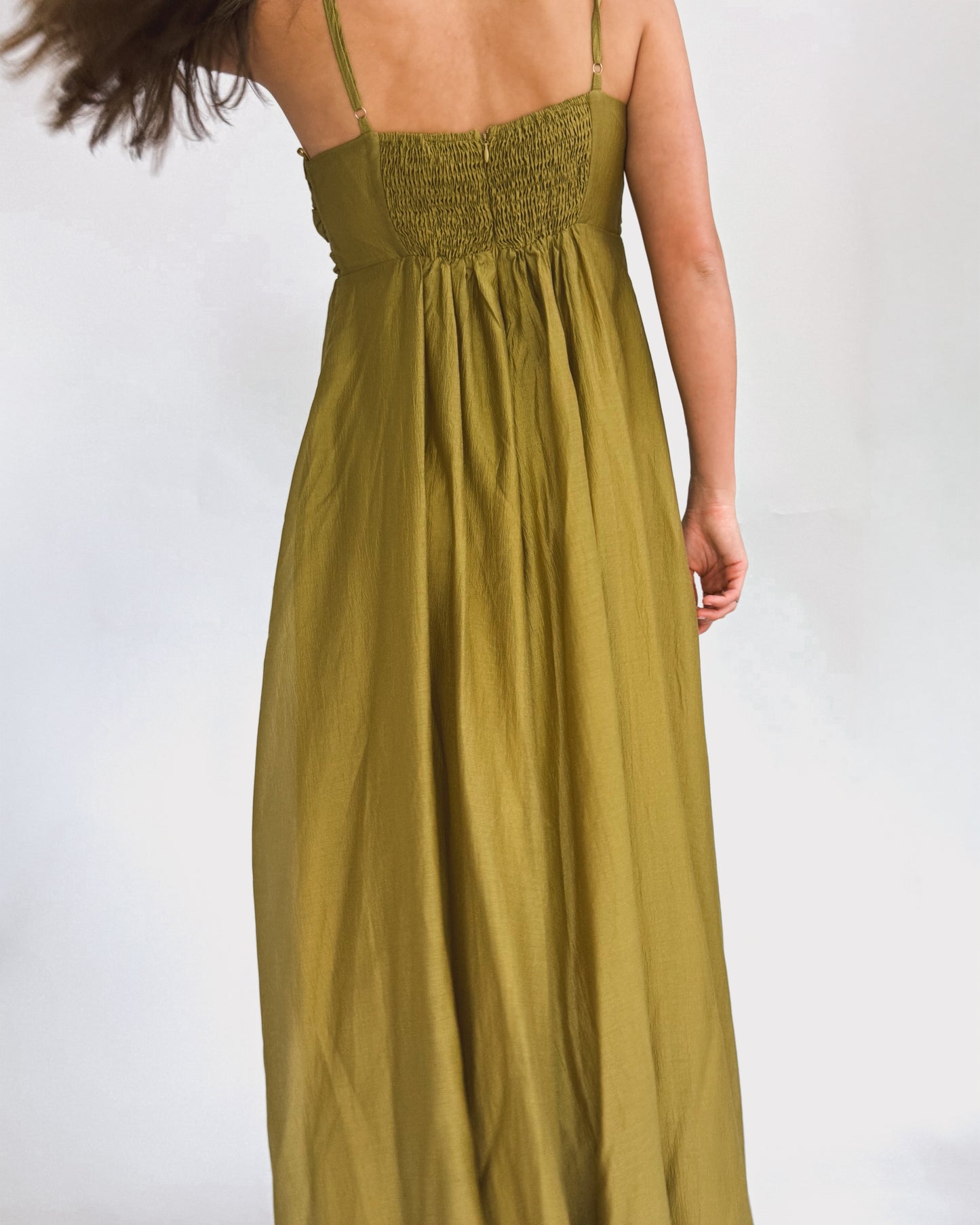 A person wearing a long, olive green dress stands in front of a plain white background. The dress has thin straps and a gathered bust, exuding elegance.