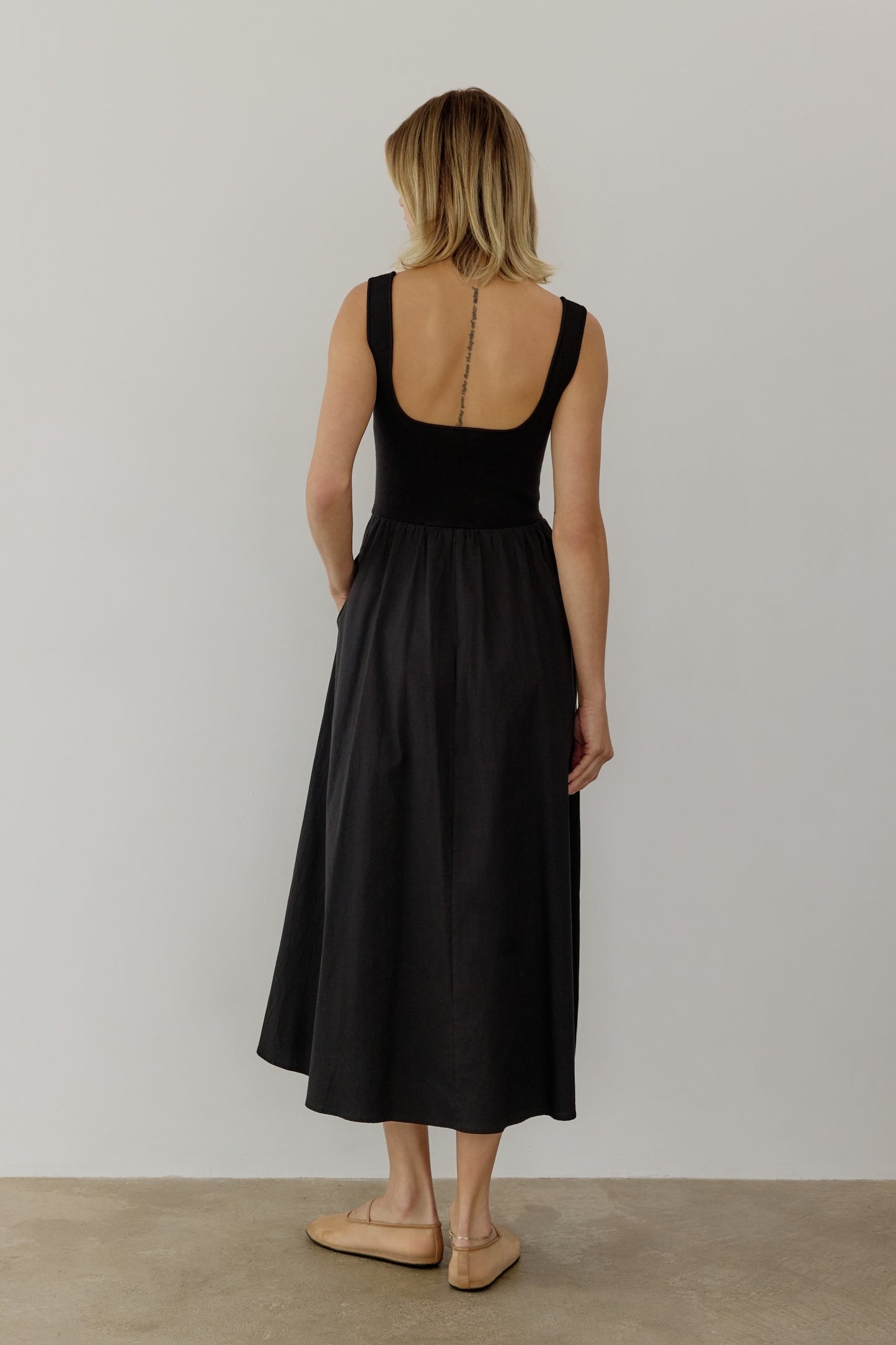 A woman in a sleeveless black midi dress stands against a plain white background. Conveying a calm and minimalist tone.