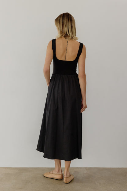 A woman in a sleeveless black midi dress stands against a plain white background. Conveying a calm and minimalist tone.