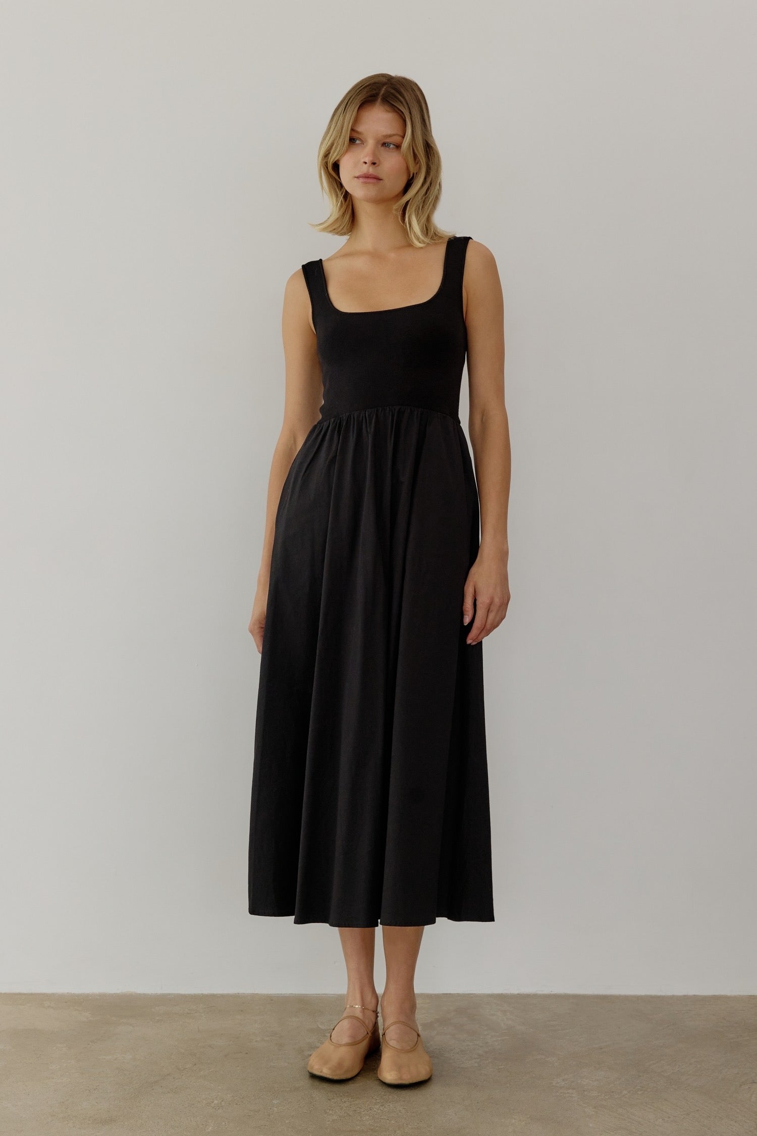 A woman in a sleeveless black midi dress stands against a plain white background. Her expression is neutral, conveying a calm and minimalist tone.
