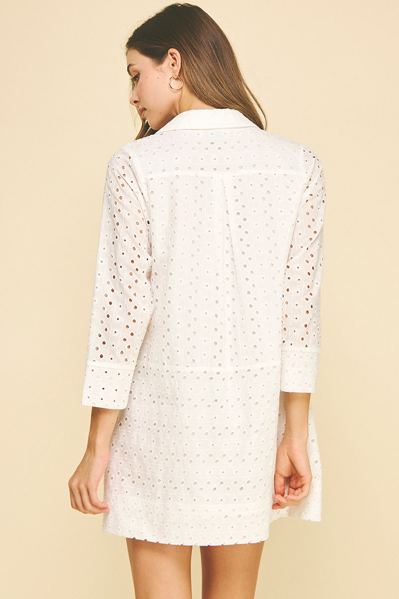 Smiling woman wearing a white perforated shirt dress with buttons, standing against a light beige background. She exudes a relaxed, confident vibe.