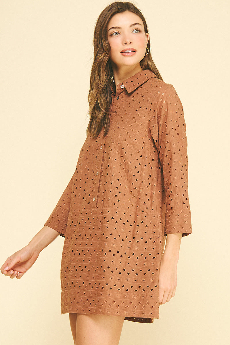 Smiling woman wearing a rust orange perforated shirt dress with buttons, standing against a light beige background. She exudes a relaxed, confident vibe.