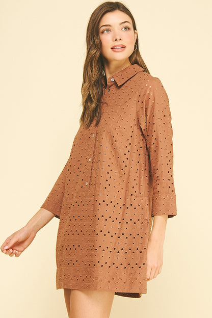 Smiling woman wearing a rust orange perforated shirt dress with buttons, standing against a light beige background. She exudes a relaxed, confident vibe.