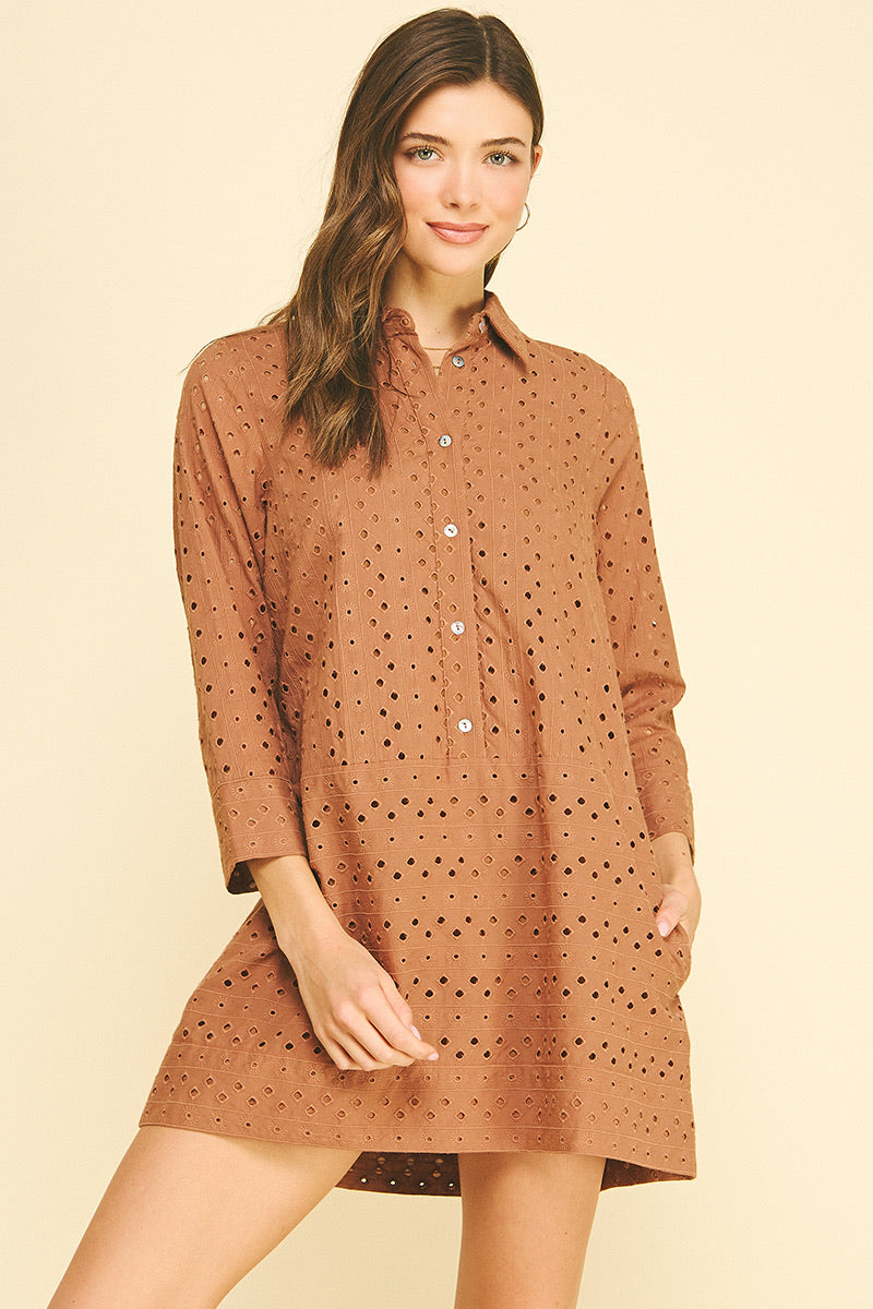 Smiling woman wearing a rust orange perforated shirt dress with buttons, standing against a light beige background. She exudes a relaxed, confident vibe.