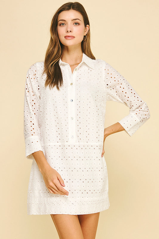 Smiling woman wearing a white perforated shirt dress with buttons, standing against a light beige background. She exudes a relaxed, confident vibe.