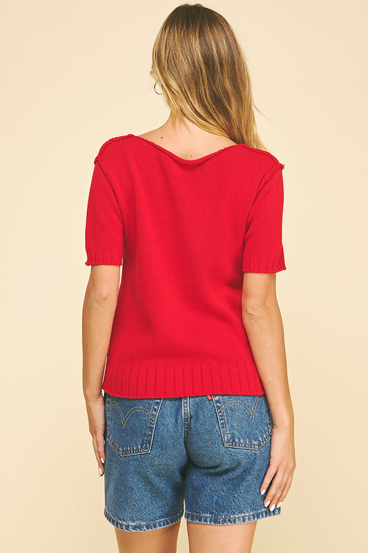 A person with long blonde hair wearing a bright red short-sleeve sweater and blue jeans shorts, set against a plain beige background.