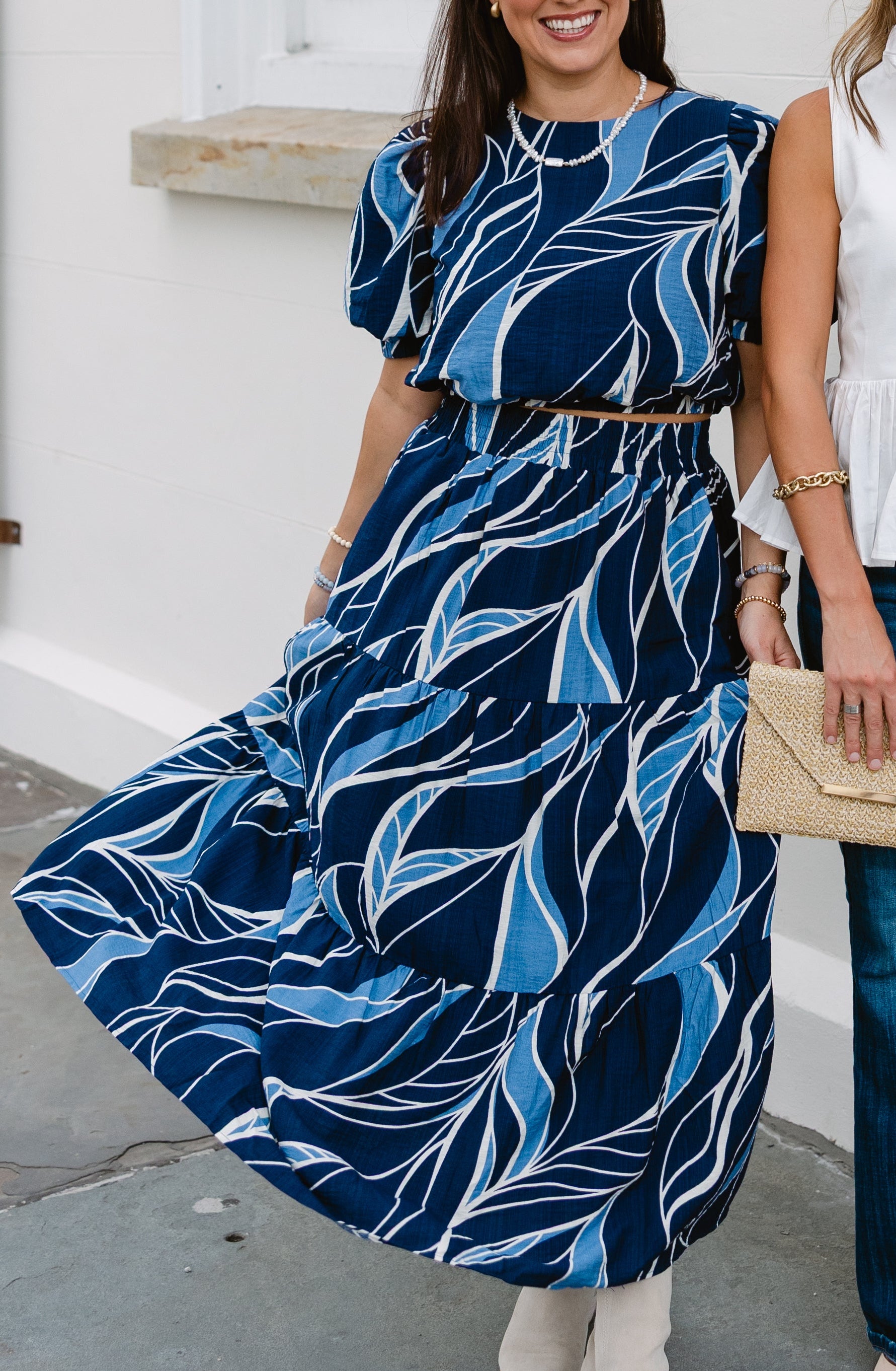Woman wearing a vibrant blue dress with a flowing abstract leaf pattern stands smiling. She holds a straw clutch, exuding a cheerful, summery vibe.