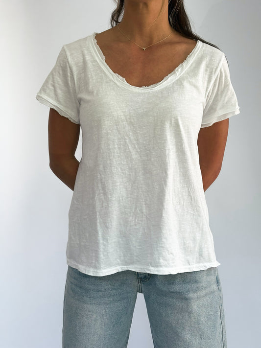 Woman wearing a casual white V-neck T-shirt and light blue jeans. Her arms are behind her back, and she wears a delicate necklace. The setting is minimal and bright.
