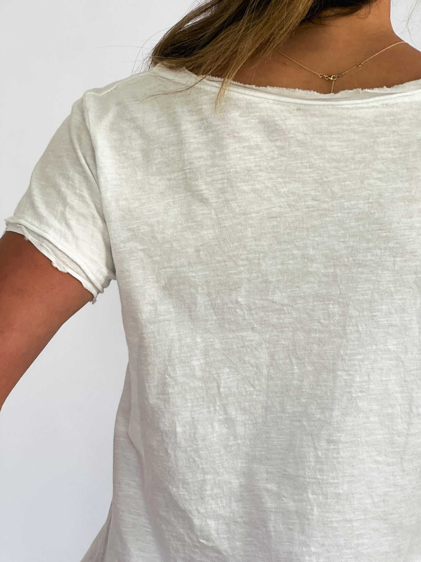 A person wearing a plain distressed white t-shirt is seen from the back against a plain background. A delicate gold necklace rests on their neck.