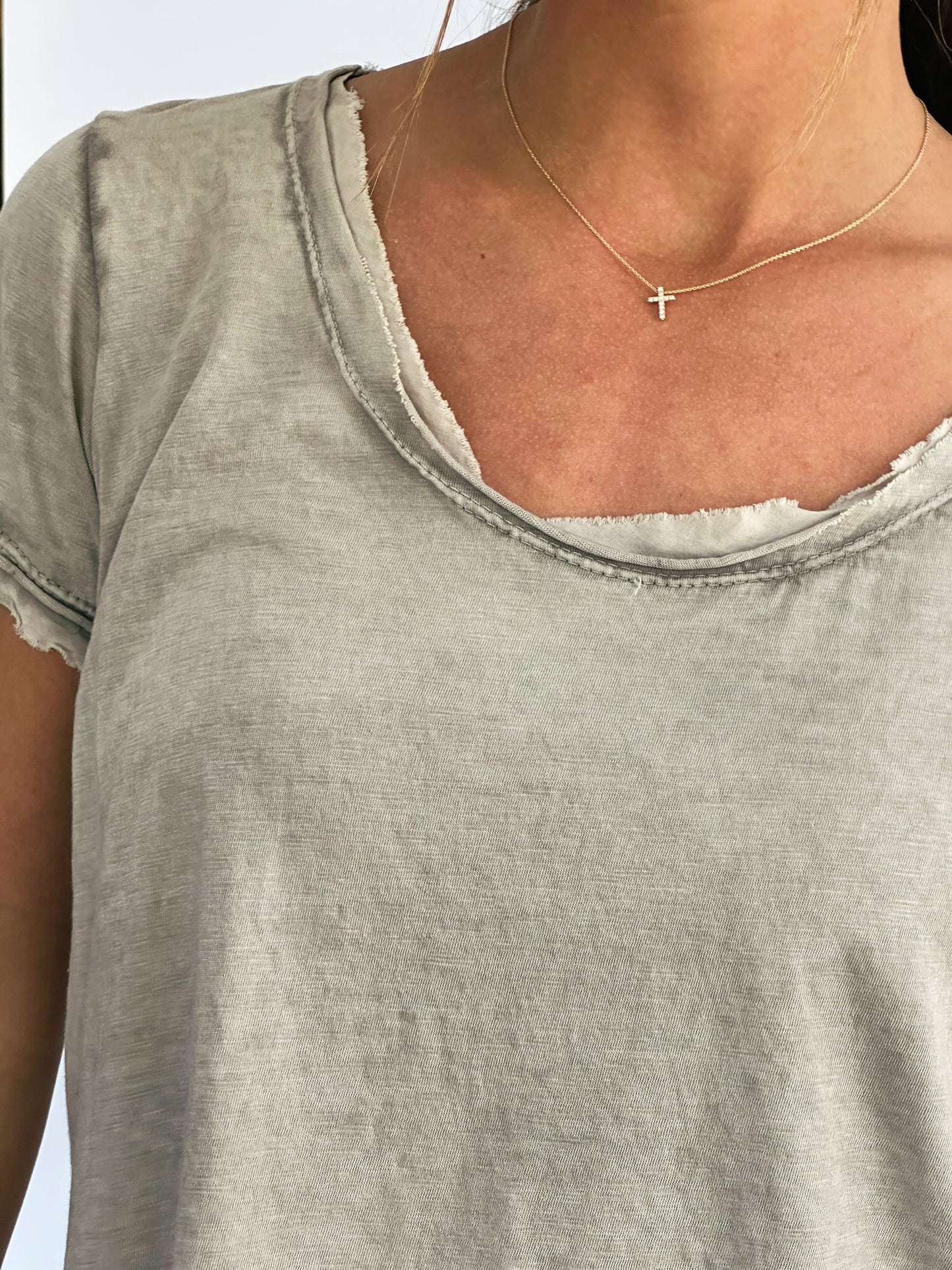 A person wearing a distressed, light taupe t-shirt with rolled sleeves and a delicate gold cross necklace. The tone is casual and understated.