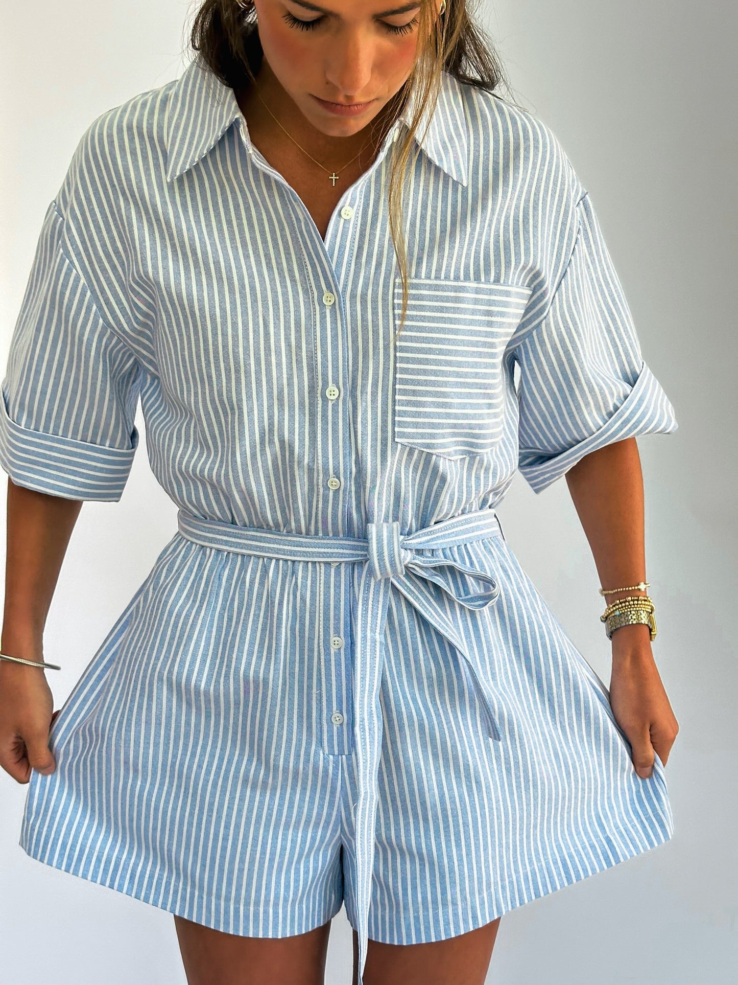 Woman wearing a light blue and white striped romper with rolled sleeves and a waist tie. 