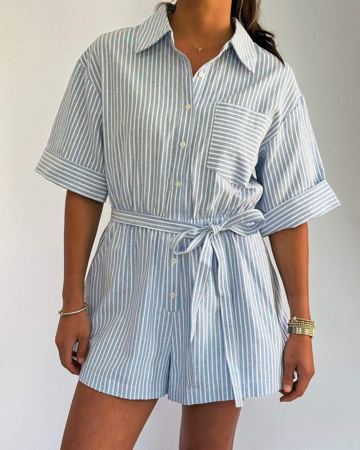 Woman wearing a light blue and white striped romper with rolled sleeves and a waist tie. 