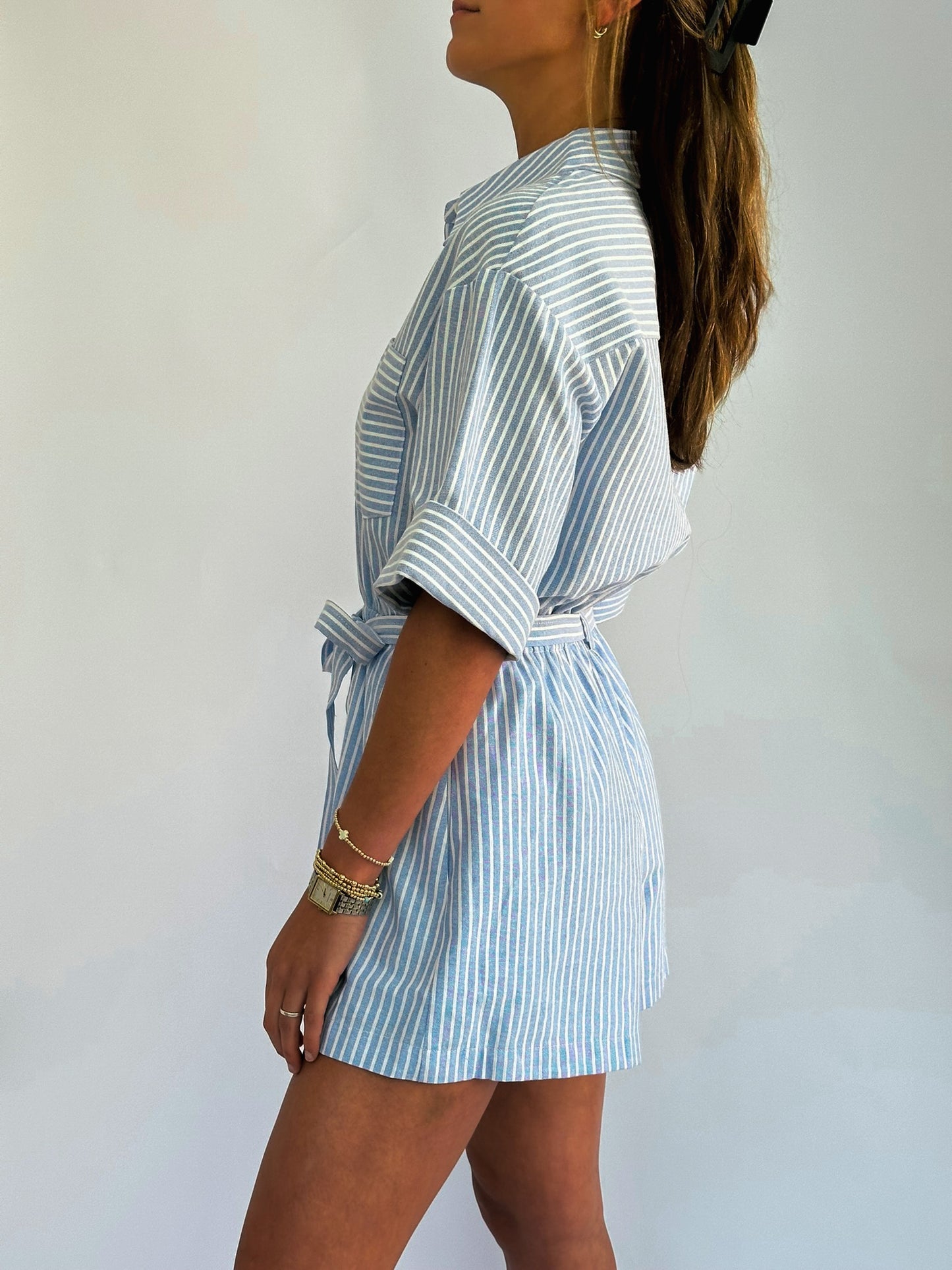 Woman in a side profile wearing a light blue and white striped romper with rolled sleeves and a waist tie. Her hair is long and features a black clip.