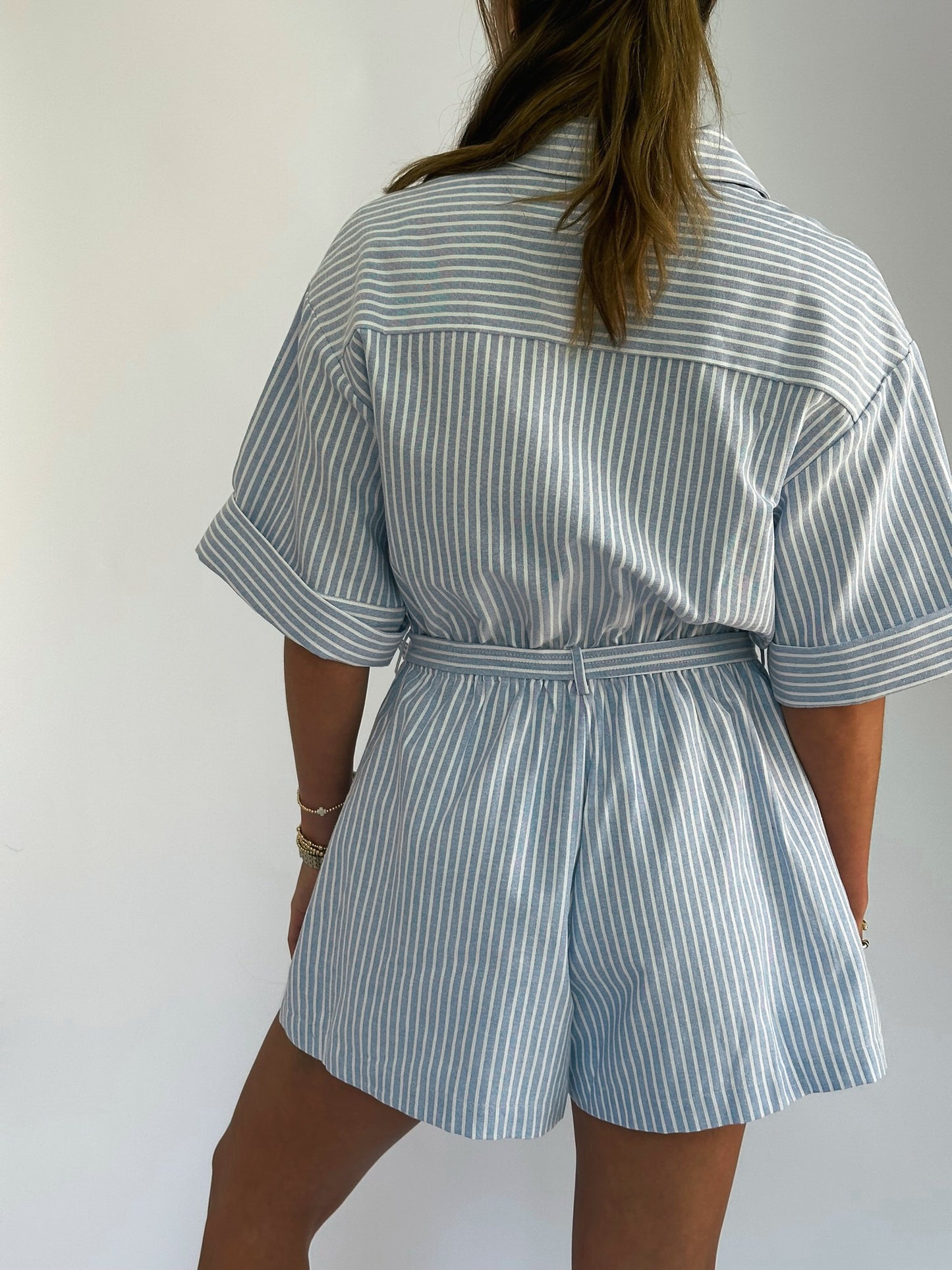 Woman wearing a light blue and white striped romper with rolled sleeves and a waist tie. 