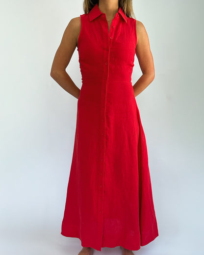 A woman wearing a sleeveless, bright red dress with a buttoned front and collared neckline stands against a white background, exuding elegance.