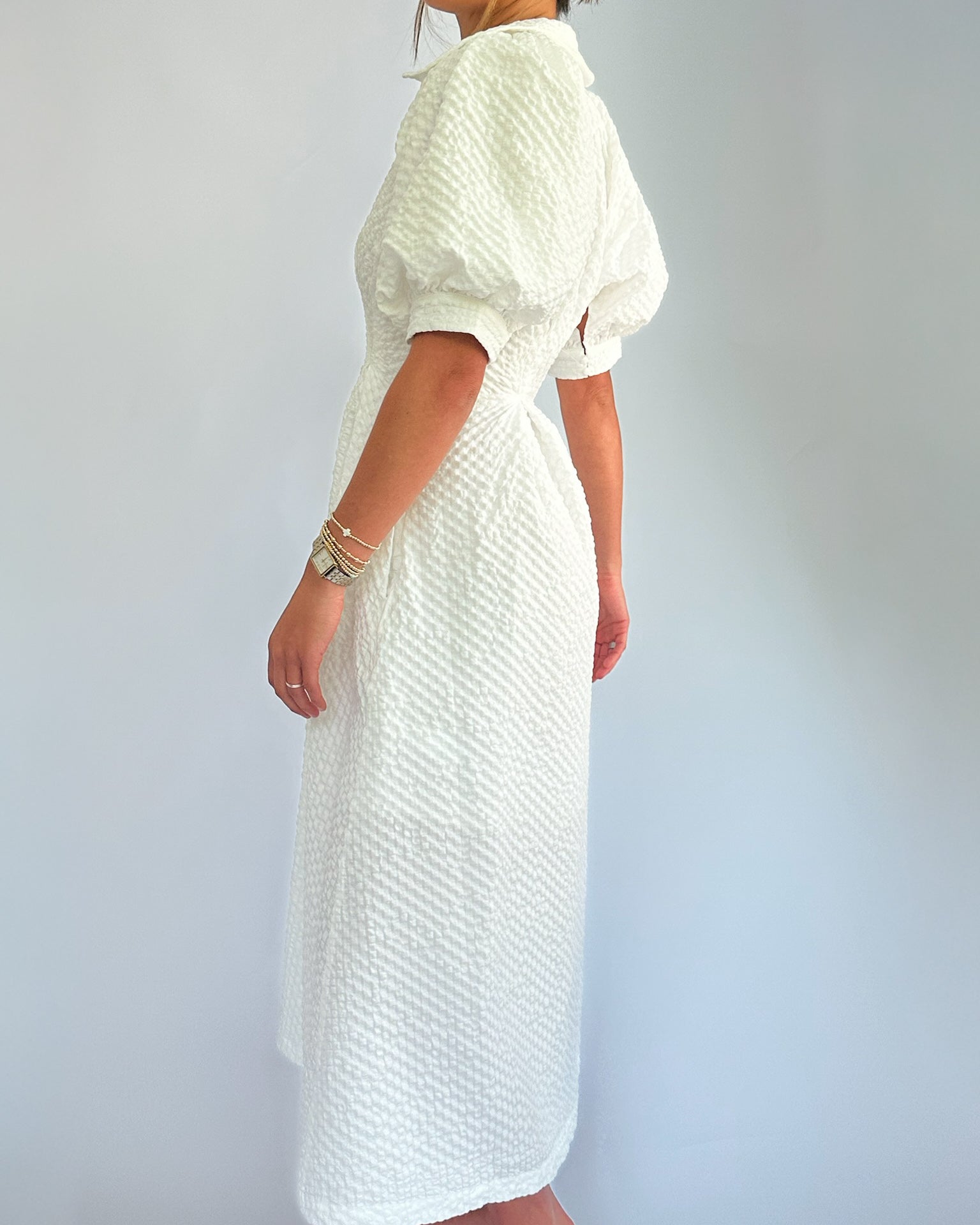 Woman in a textured white dress with puff sleeves, standing sideways. She wears gold bracelets, conveying elegance against a neutral background.
