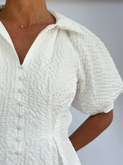 Close-up of a person wearing a textured white dress with buttons and puffed sleeves. A delicate star necklace adorns their neck, exuding elegance.