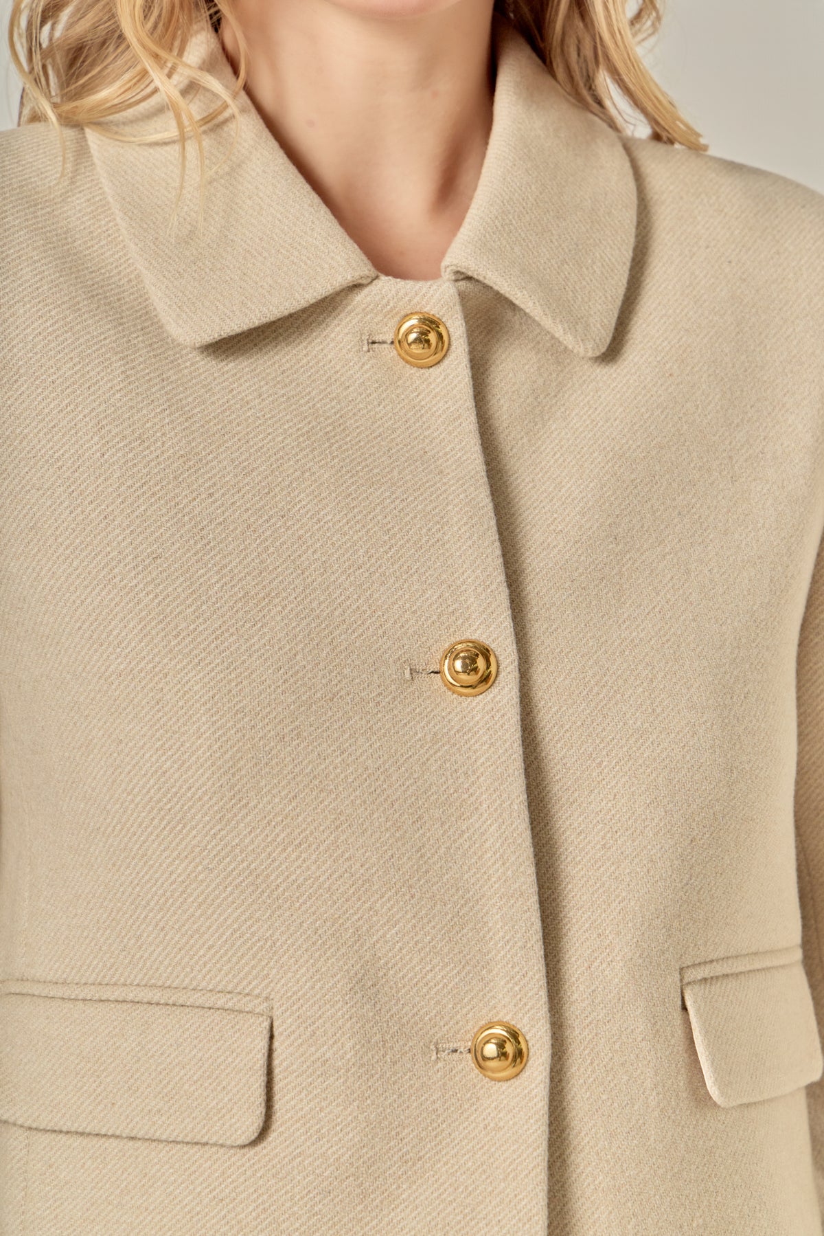 A woman with wavy blonde hair wears a beige buttoned jacket and matching shorts. 