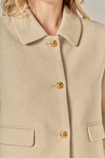 A woman with wavy blonde hair wears a beige buttoned jacket and matching shorts. 