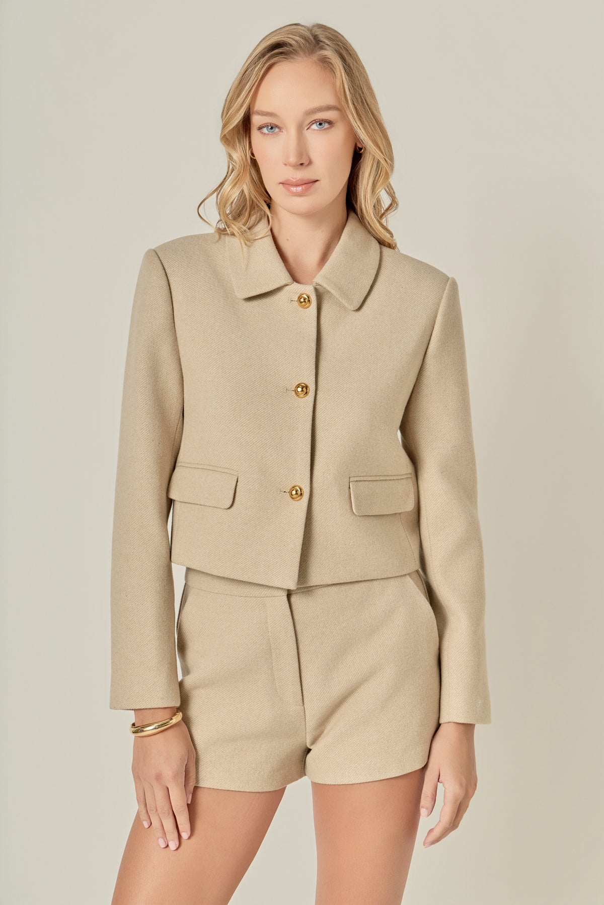 A woman with wavy blonde hair wears a beige buttoned jacket and matching shorts. She looks at the camera with a calm expression, conveying elegance.