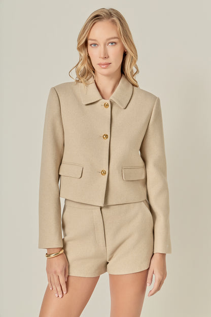A woman with wavy blonde hair wears a beige buttoned jacket and matching shorts. She looks at the camera with a calm expression, conveying elegance.