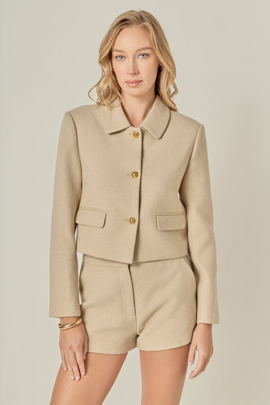 A woman with wavy blonde hair wears a beige buttoned jacket and matching shorts. She looks at the camera with a calm expression, conveying elegance.