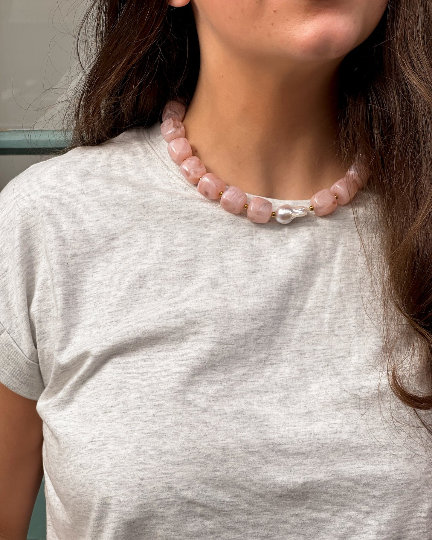 A woman in a light grey t-shirt wears a chunky pink bead necklace. Her long dark hair frames a serene, slightly smiling expression.