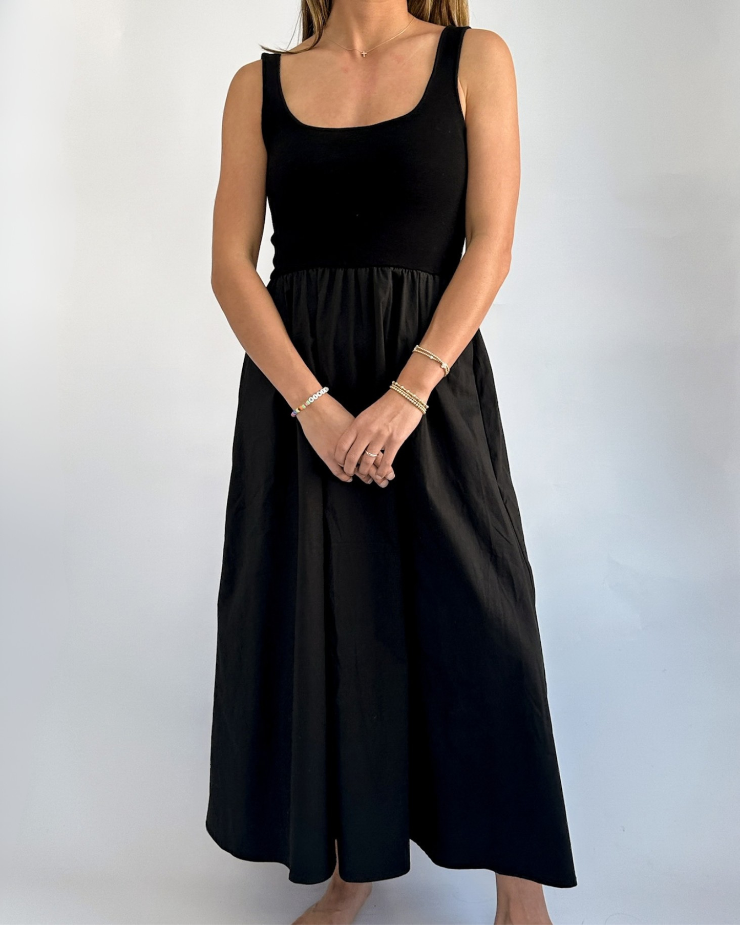 A woman stands against a plain light background, wearing a black sleeveless dress. Her arms are crossed lightly in front, showcasing her bracelets.